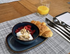 Breakfast bowl containing fresh fruit and yoghurt with a croissant on the side, along with a half glass of orange juice